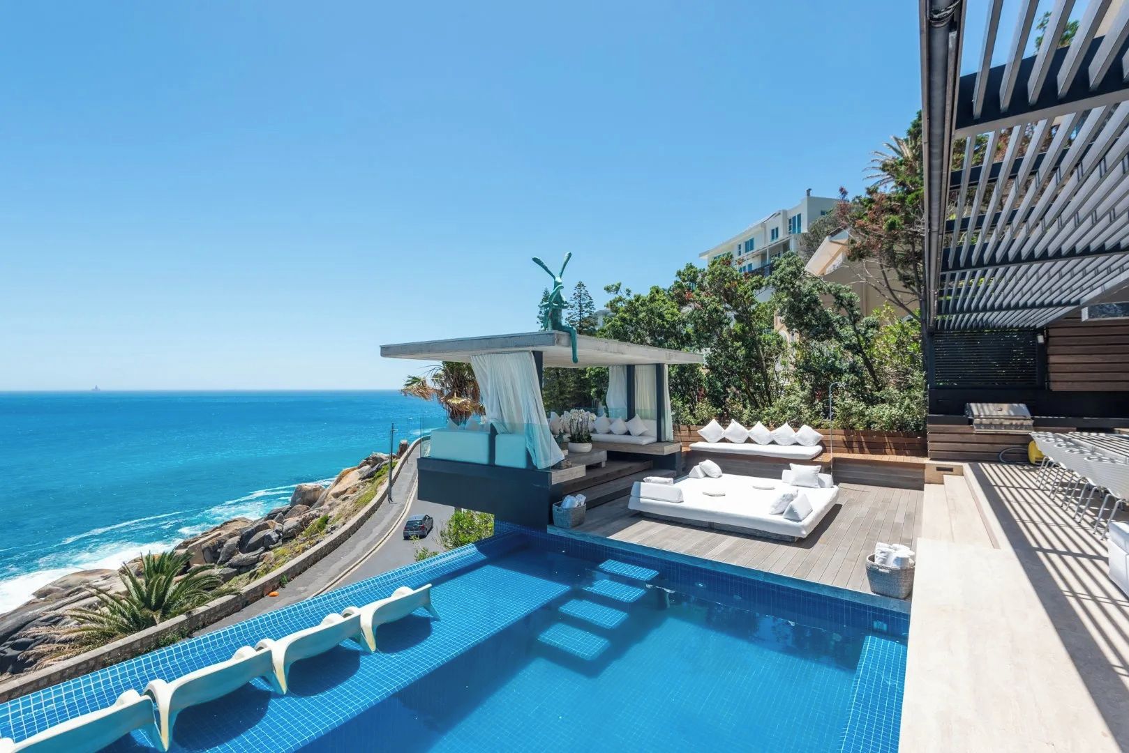 A rooftop infinity pool with a view of the ocean, outdoor seating with cushions, and a modern architectural design.