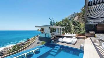 A rooftop infinity pool with a view of the ocean, outdoor seating with cushions, and a modern architectural design.
