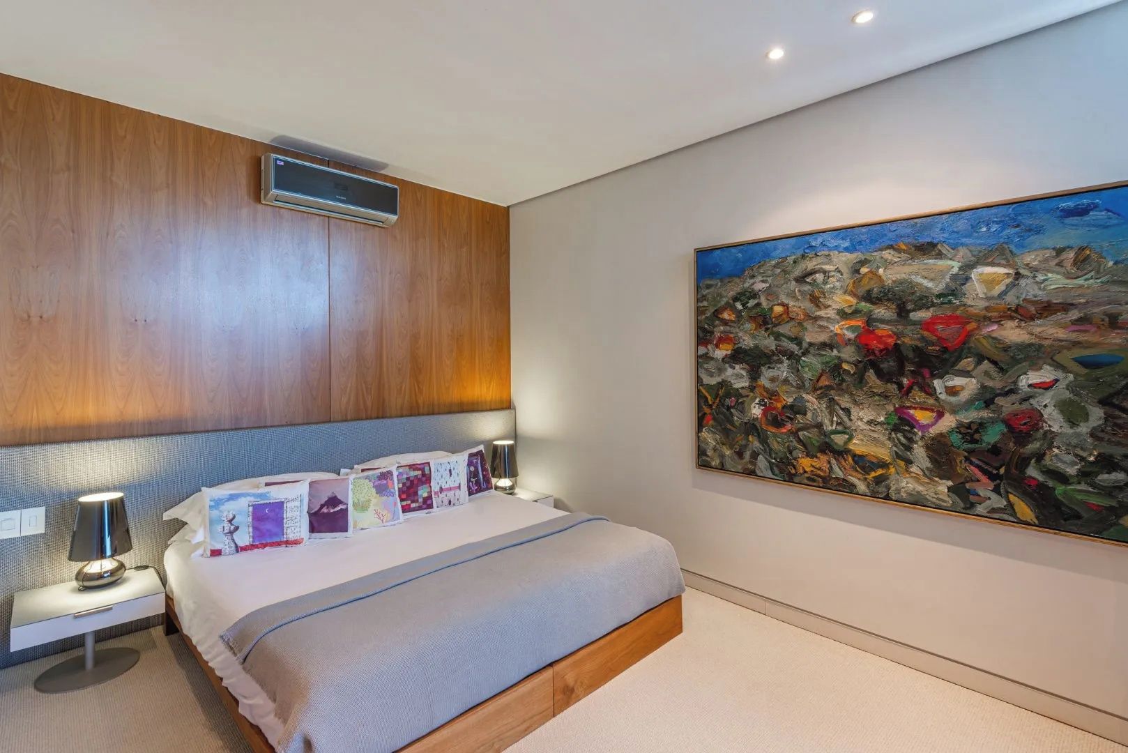 A bedroom with a bed, a nightstand, a lamp, a painting, and a wall-mounted air conditioner.