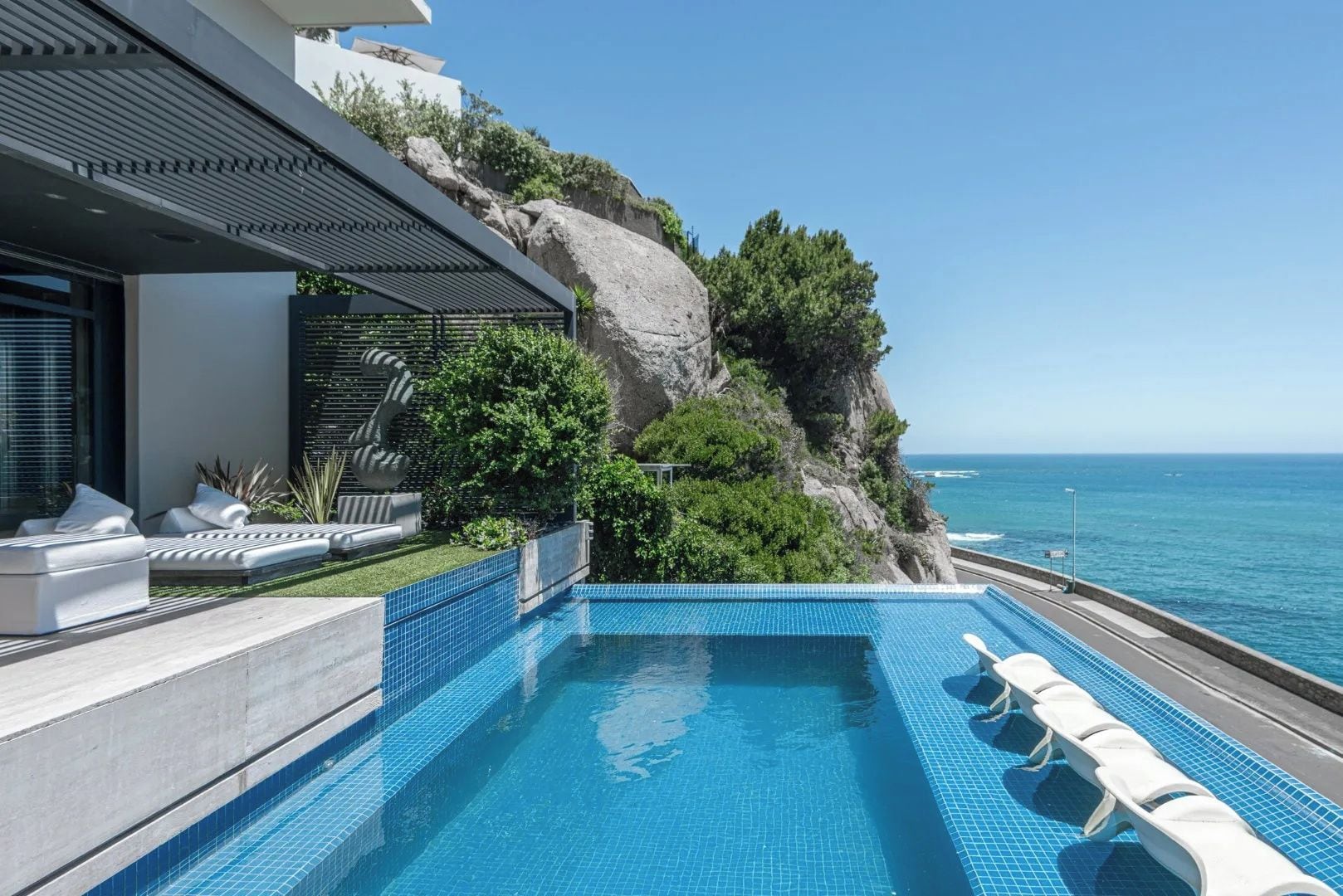 A modern outdoor swimming pool with a view of the ocean, a rocky cliff, and a lounge area with white cushions.
