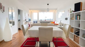 A modern living space with a dining area, a sofa, a bookshelf, and a view of the outdoors.