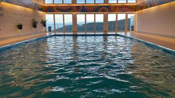 Ein Hallenbad mit klarem Wasser, großen Fenstern und einem farbenfrohen, abstrakten Wandgemälde.
