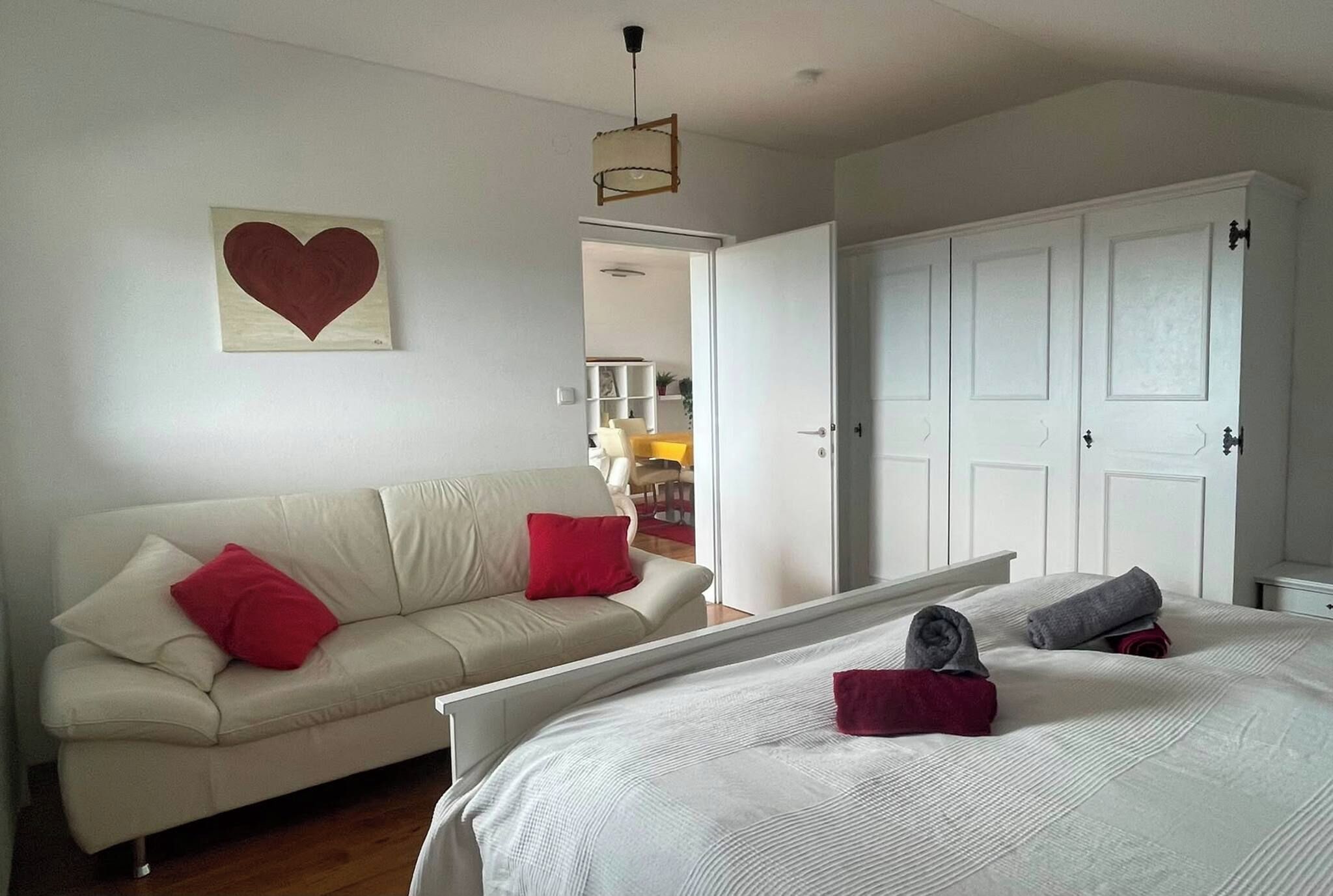 Une chambre à coucher avec un lit, un canapé blanc orné de coussins rouges, une décoration en forme de cœur accrochée au mur et une porte donnant sur une autre pièce.