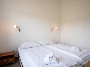 Een enkel bed met witte lakens en twee kussens, een houten nachtkastje met een lade, en twee wandlampen.
