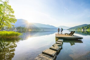 Een rustig meer met een houten steiger die zich in het water uitstrekt, omringd door groen en bergen op de achtergrond.