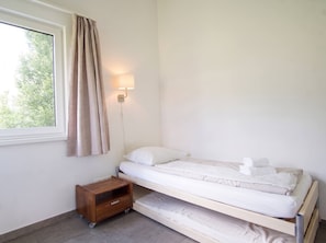 Een eenpersoonsbed met een houten nachtkastje en een wandlamp in een kamer met een raam en gordijnen.