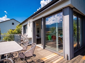 Een modern huis met een houten terras, een eethoek met stoelen en een tafel, en grote schuifdeuren die leiden naar een binnenruimte.