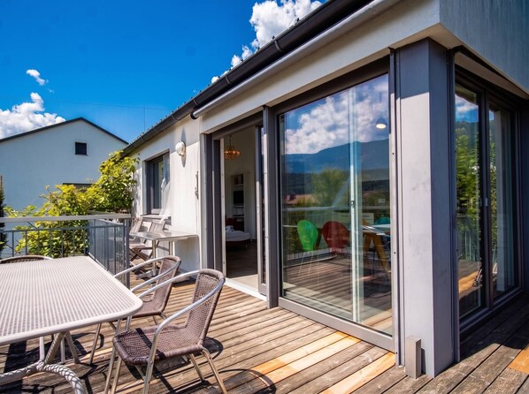 Een modern huis met een houten terras, een eethoek met stoelen en een tafel, en grote schuifdeuren die leiden naar een binnenruimte.