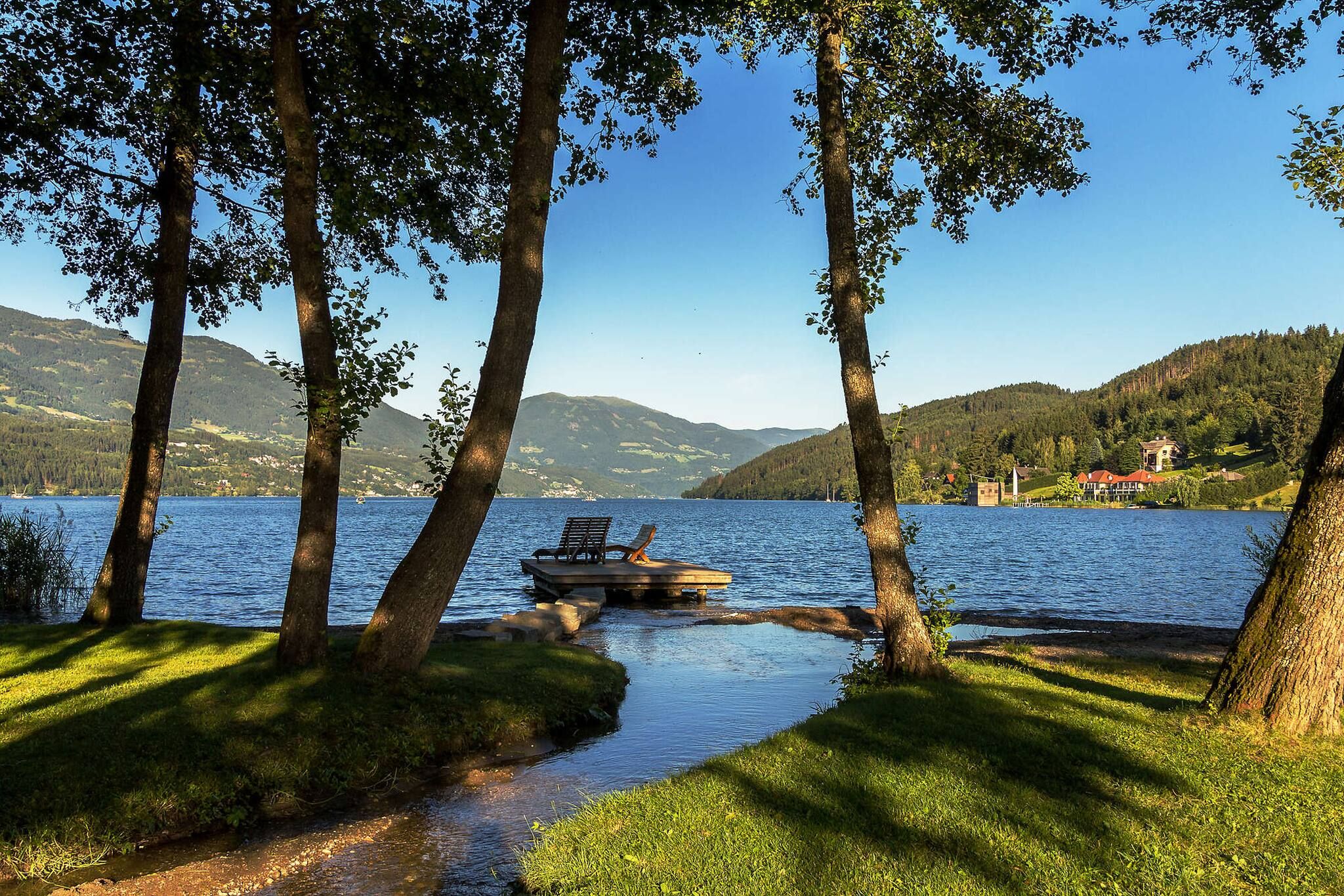 A lakeside view with a dock, trees, and a distant house.