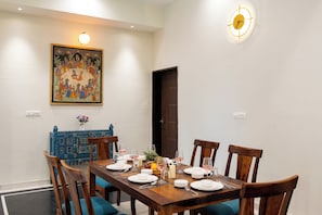 A dining room with a wooden table set for four, surrounded by chairs, a painting on the wall, and a clock. - Alaya Stays Leora Amar Villa 6BHK (Udaipur)
