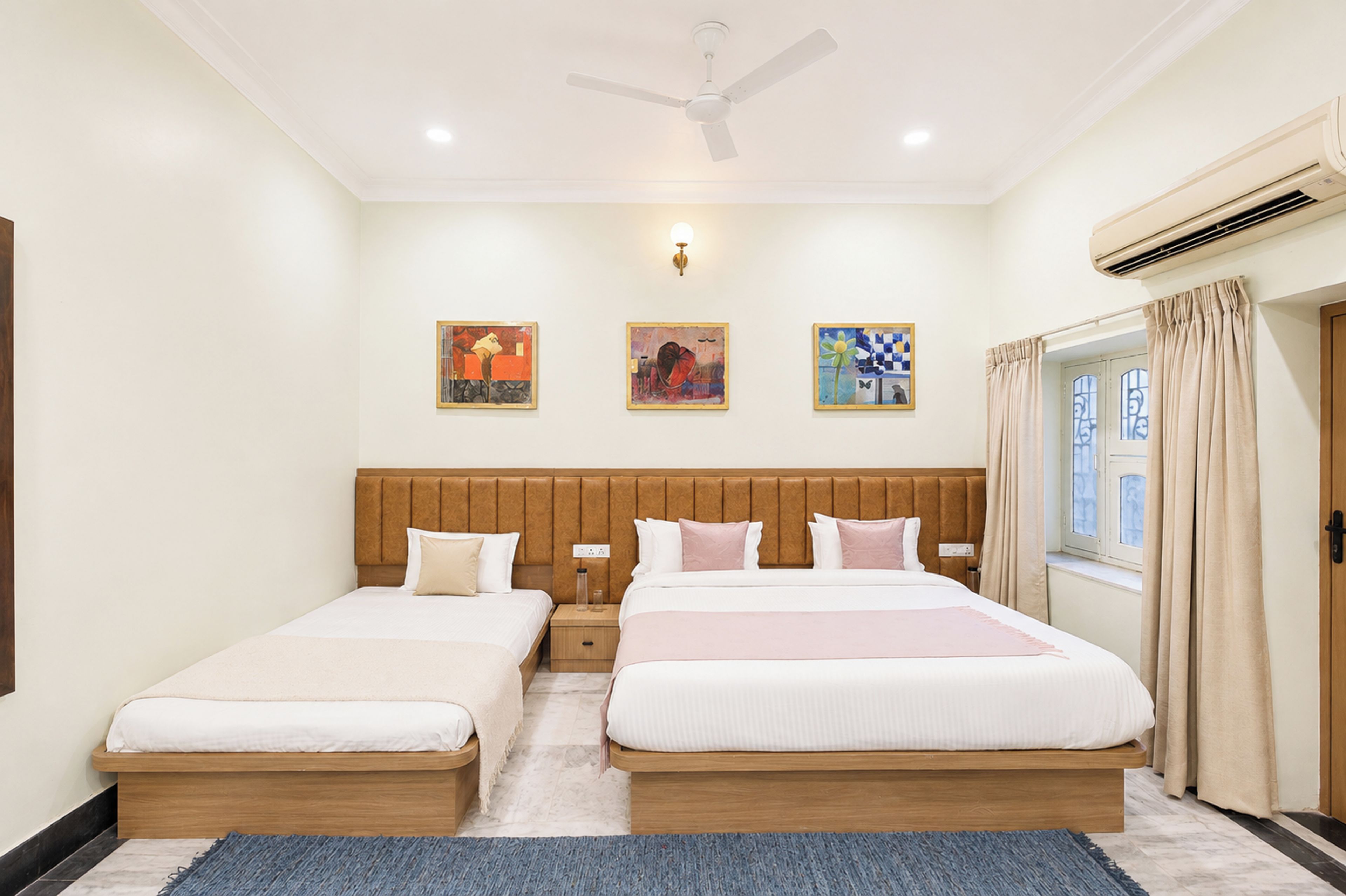 A hotel room with two beds, a ceiling fan, wall-mounted artwork, and an air conditioning unit.