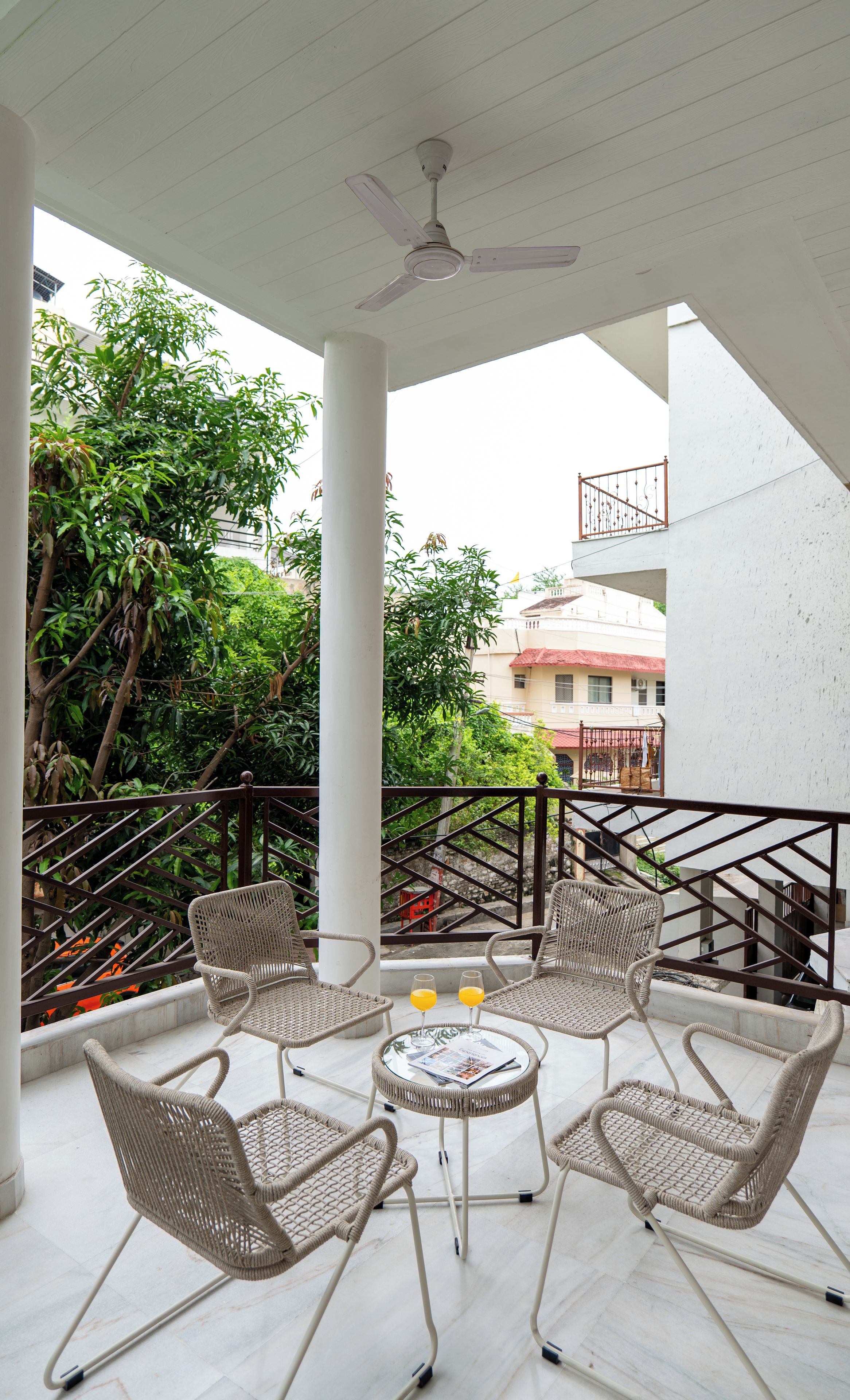 A covered outdoor seating area with wicker chairs and a table, a ceiling fan, and a view of greenery and buildings in the background.
