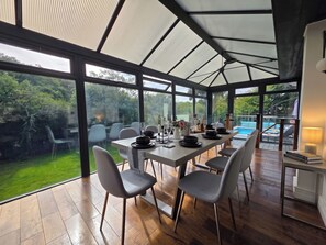 A conservatory with a dining table set for six, surrounded by chairs, with a view of a swimming pool and greenery outside. - House Sleeps 11 With Outdoor Pool (Lymington)