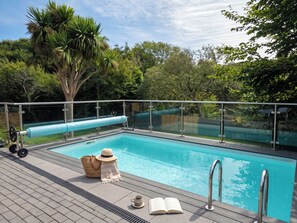 A swimming pool with a glass barrier, a lounge area with a hat and book, and a tree in the background. - House Sleeps 11 With Outdoor Pool (Lymington)