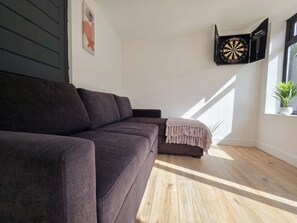 A modern living room with a dark-colored sofa, a dartboard, and a window with natural light. - House Sleeps 11 With Outdoor Pool (Lymington)