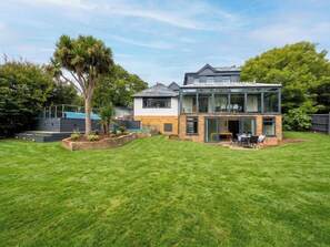 A modern two-story house with a glass conservatory, a well-maintained lawn, and a variety of trees. - House Sleeps 11 With Outdoor Pool (Lymington)