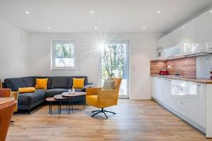 Ein modernes Wohnzimmer mit einer Couch, einem Couchtisch und einem Sessel, eine Küche mit weißen Schränken und einer Holzarbeitsplatte sowie große Fenster mit Tageslicht.
