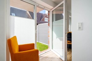 A room with a sliding glass door leading to a patio, a single orange chair, and a small table with a cushion. - Spiekeroog Vacation Rental (Norden)