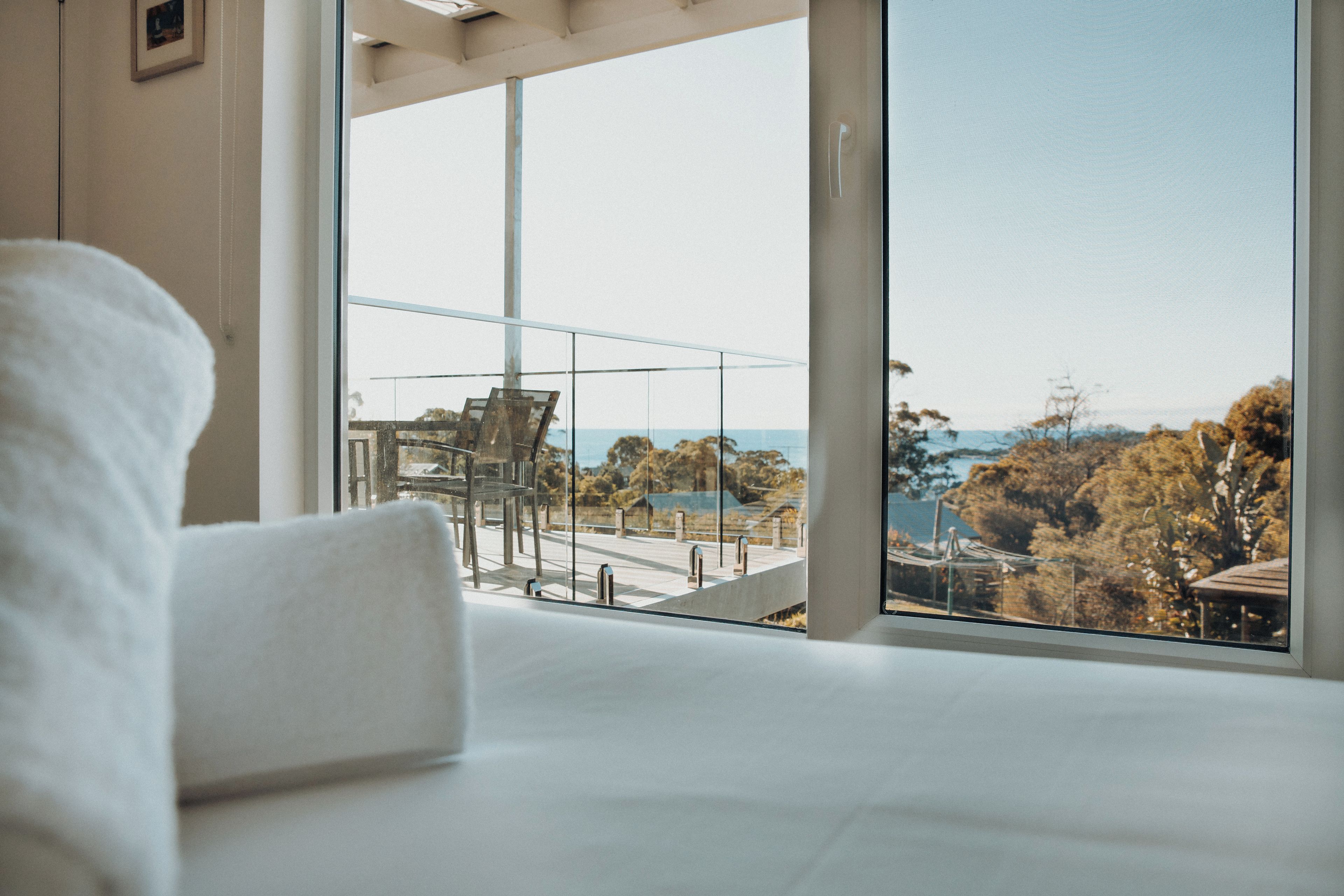 A hotel room with a large window offering a view of a balcony, outdoor furniture, and a distant landscape.