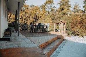 Une terrasse en bois avec des meubles d’extérieur, un espace salon couvert et une vue sur les arbres et les arbustes.