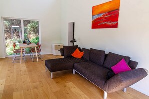 Ein modernes Wohnzimmer mit einer Eckcouch, einem Esstisch mit Stühlen und einem großen Fenster mit Blick ins Grüne.