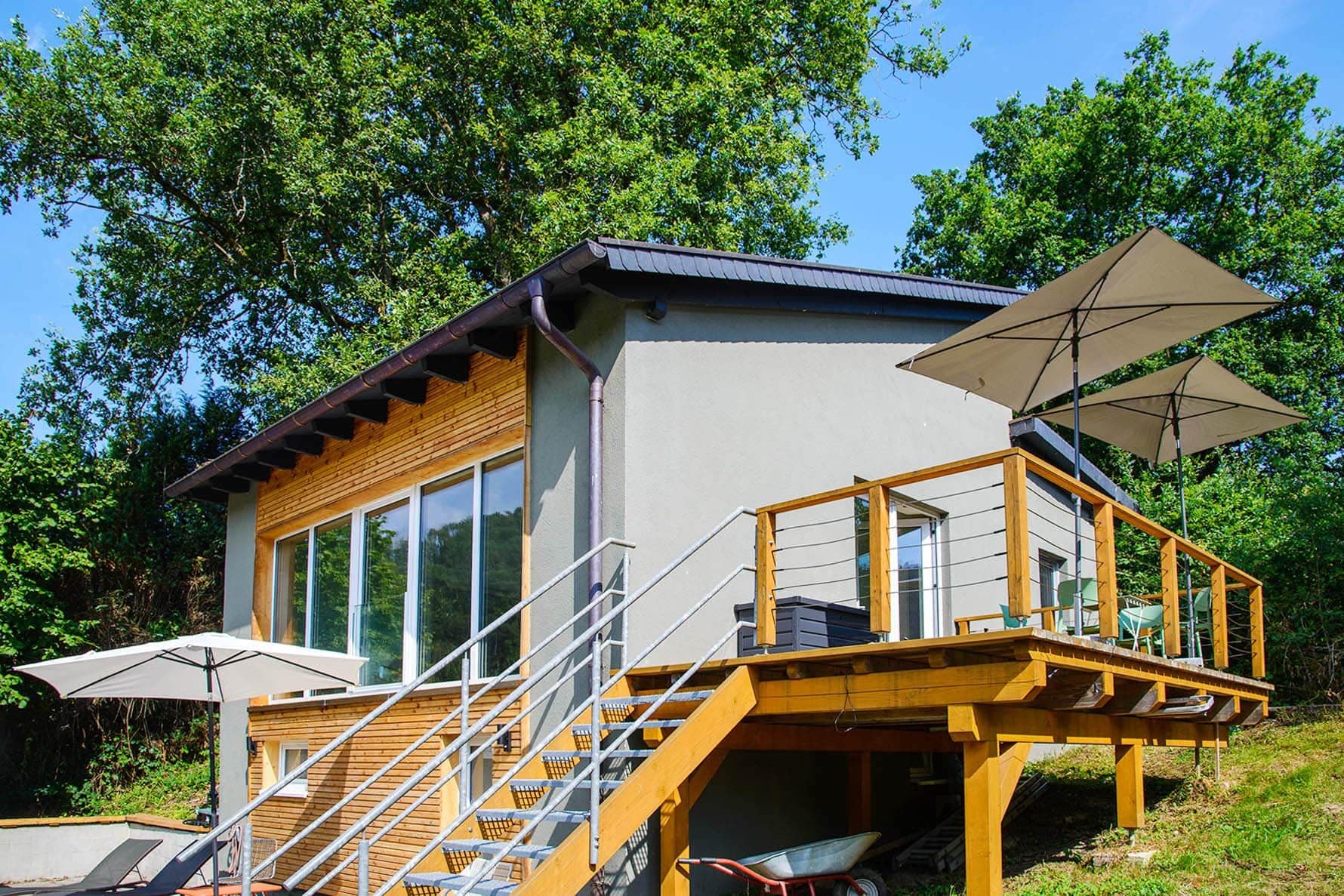 A modern cabin with a wooden deck, outdoor seating, and a staircase leading to a balcony.