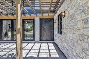 A covered outdoor area with a stone wall, a wooden ceiling, and a door leading to a glass entrance. - New Ultra Luxury Retreat minutes from Arcadia! (Phoenix)