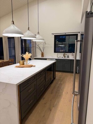 Une cuisine moderne dotée d’un grand îlot central, de luminaires suspendus et de meubles de rangement aux couleurs sombres.