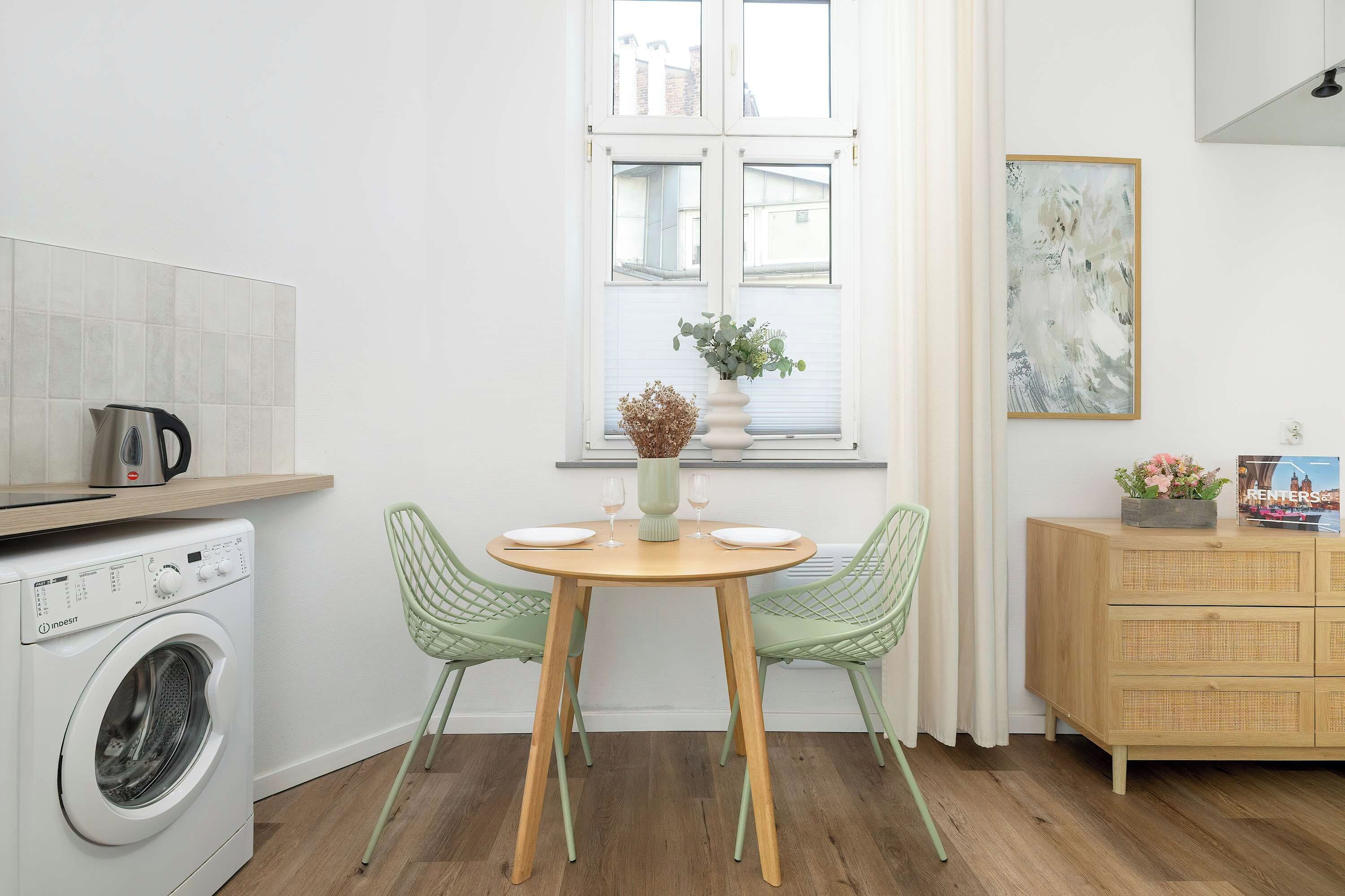 A compact kitchen with a washing machine, a dining table with chairs, a window with blinds, and a cabinet with a framed picture.