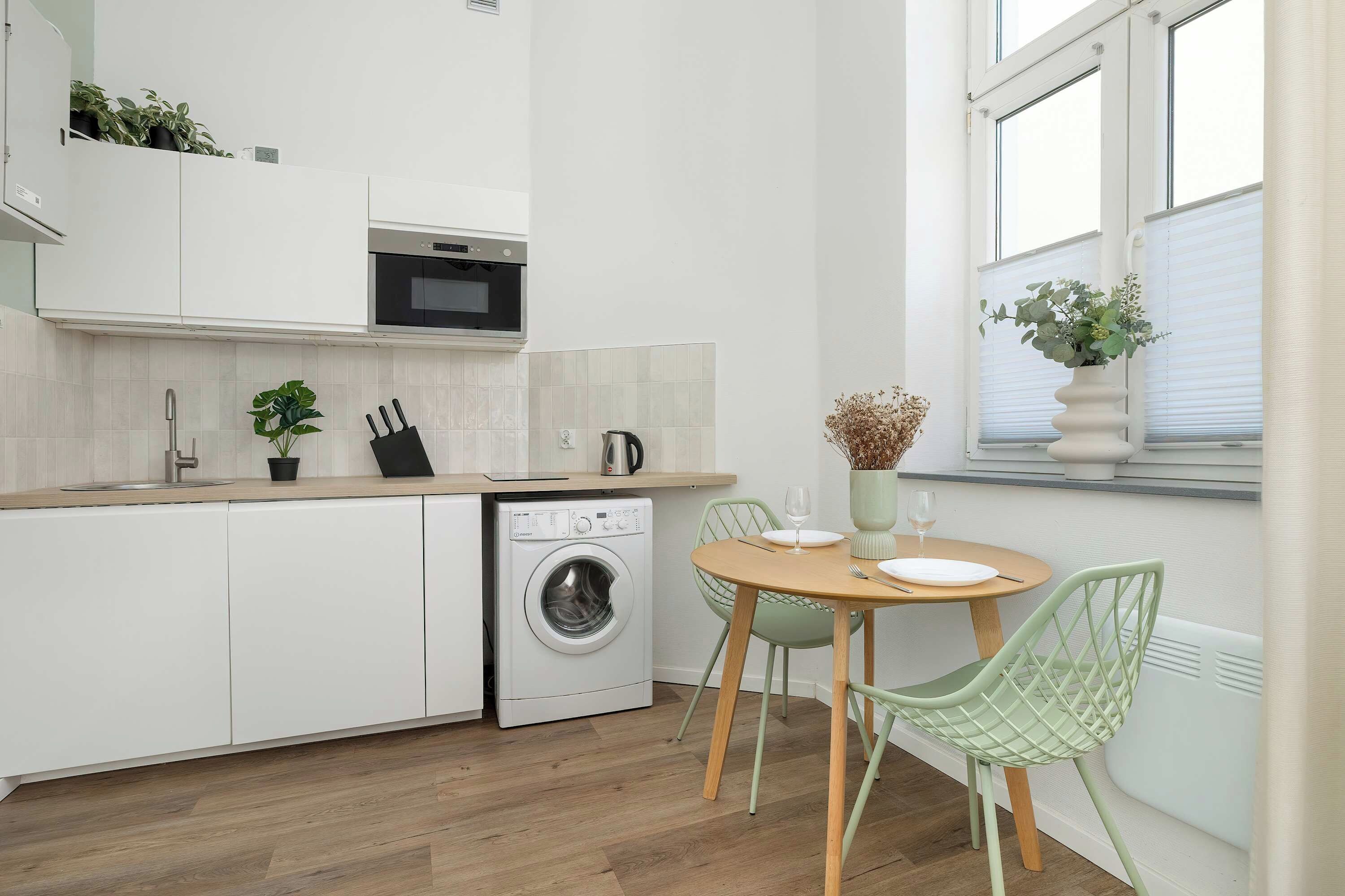 Een moderne keuken met een ingebouwde wasmachine, een eethoek met een tafel en stoelen, en een raam met een plant.