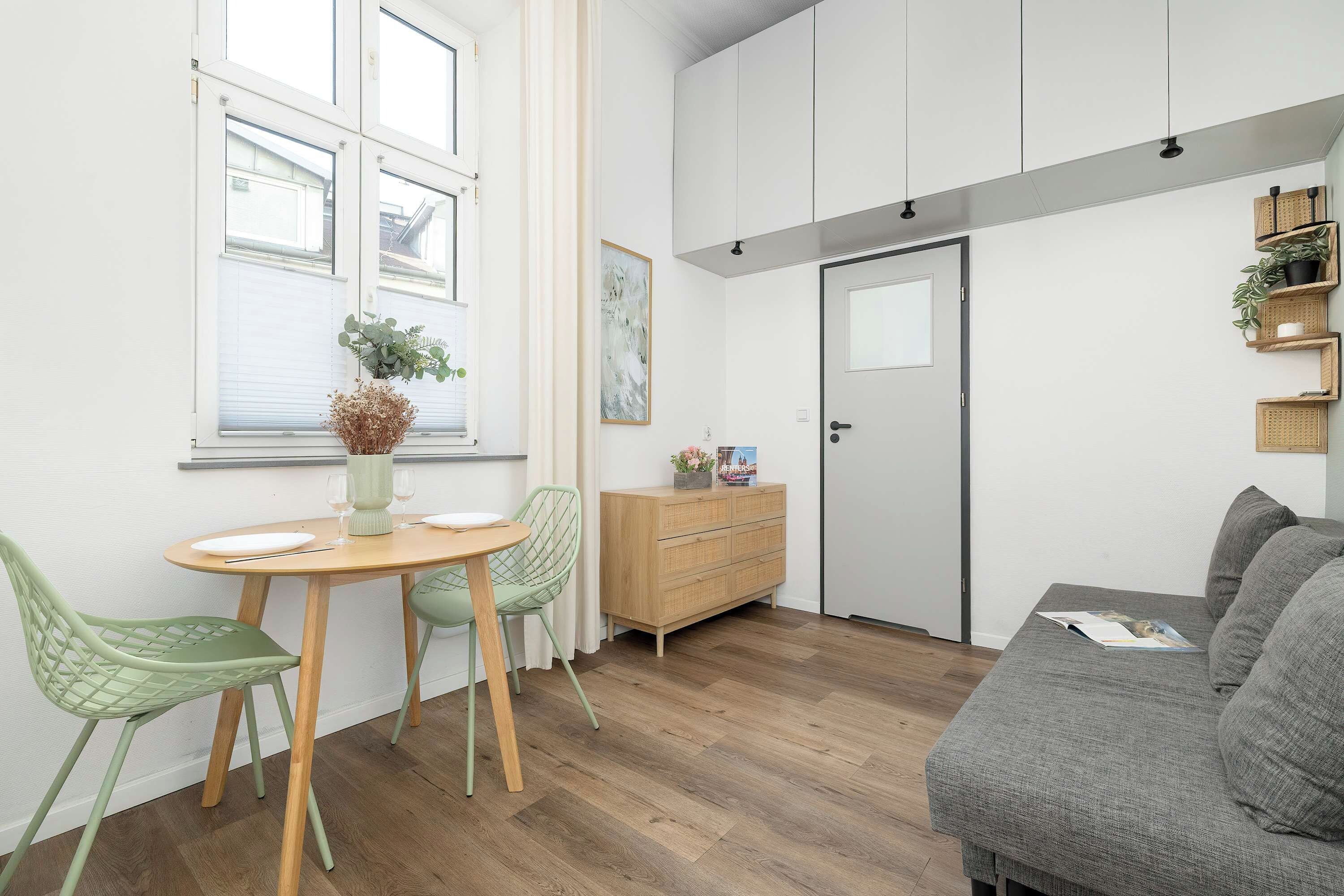 A modern living room with a wooden floor, a grey sofa, a dining table with chairs, a wooden cabinet, and a door.
