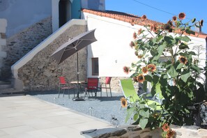 A building with a stone facade, a tiled roof, and a staircase leading to an upper level. There is a patio area with outdoor seating and a plant with sunflowers. - Cottage 'L'entredeux' with Shared Terrace, Shared Garden and Wi-Fi (Saint-Viaud)