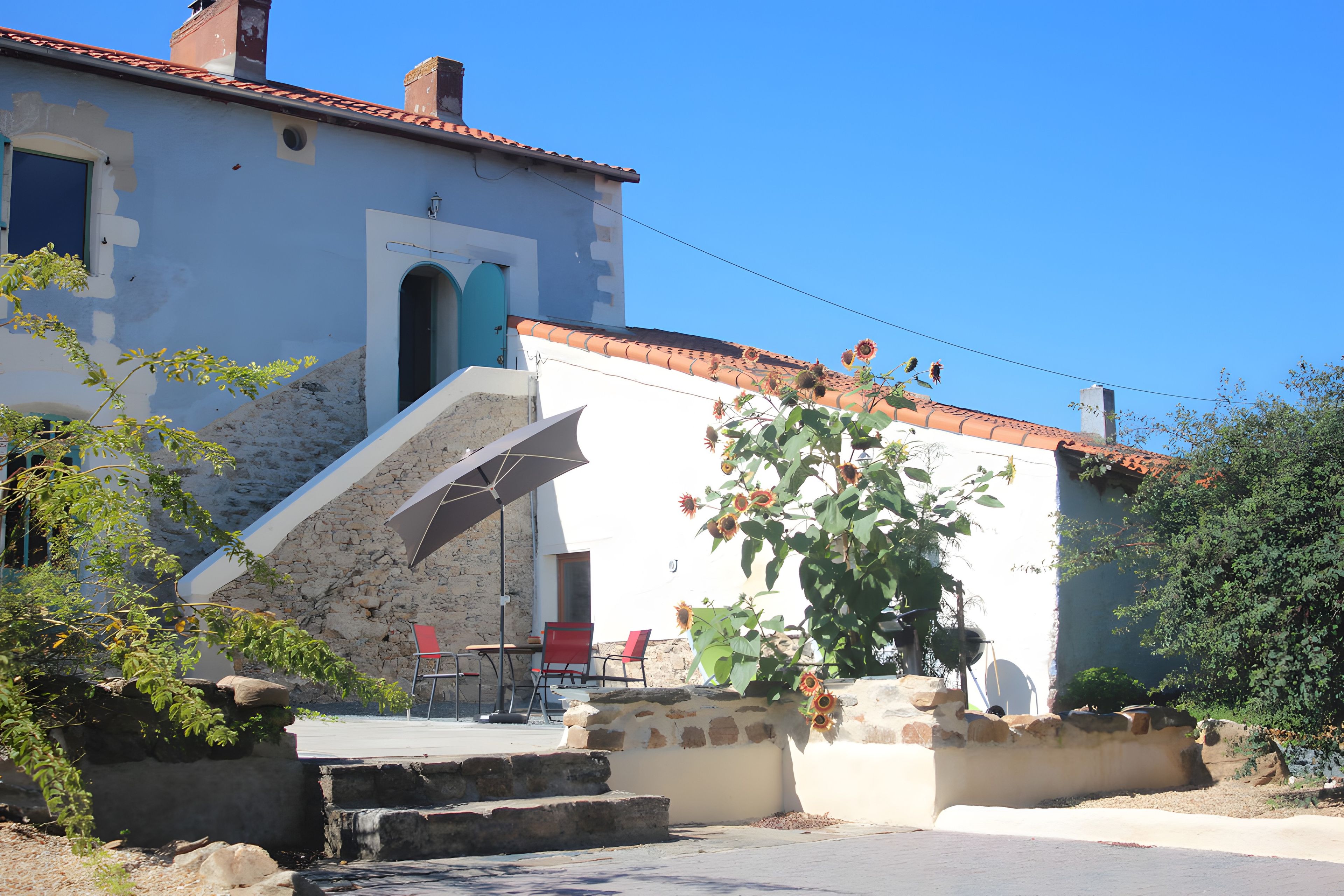 Un edificio con una scala in pietra che conduce a una porta, una piccola zona esterna con tavolo e sedie, e un giardino con girasoli.