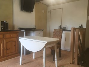 A compact kitchen with a white table, wooden cabinets, a built-in dishwasher, and a television mounted on the wall. - Cottage 'L'entredeux' with Shared Terrace, Shared Garden and Wi-Fi (Saint-Viaud)