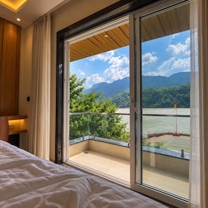 A hotel room with a large sliding glass door leading to a balcony with a view of a lake and mountains. - Shree Rajneesh Osho Ashram (Rishikesh)
