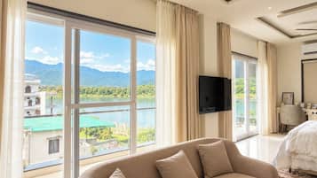 A hotel room with a large window offering a view of a lake and mountains, a flat-screen TV mounted on the wall, and a sofa with cushions.