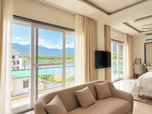 A hotel room with a large window offering a view of a lake and mountains, a flat-screen TV mounted on the wall, and a sofa with cushions. - Shree Rajneesh Osho Ashram (Rishikesh)