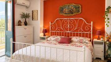 Une chambre avec un lit en métal blanc, un mur rouge orné d’un tableau encadré, une table de chevet blanche avec une lampe, et une fenêtre avec un rideau.