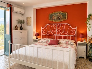 Ein Schlafzimmer mit einem weißen Metallbett, einer roten Wand mit einem gerahmten Gemälde, einem weißen Nachttisch mit Lampe und einem Fenster mit Vorhang.