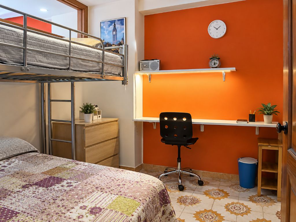 Een studentenkamer met een stapelbed, bureau, stoel en een klok aan de muur.
