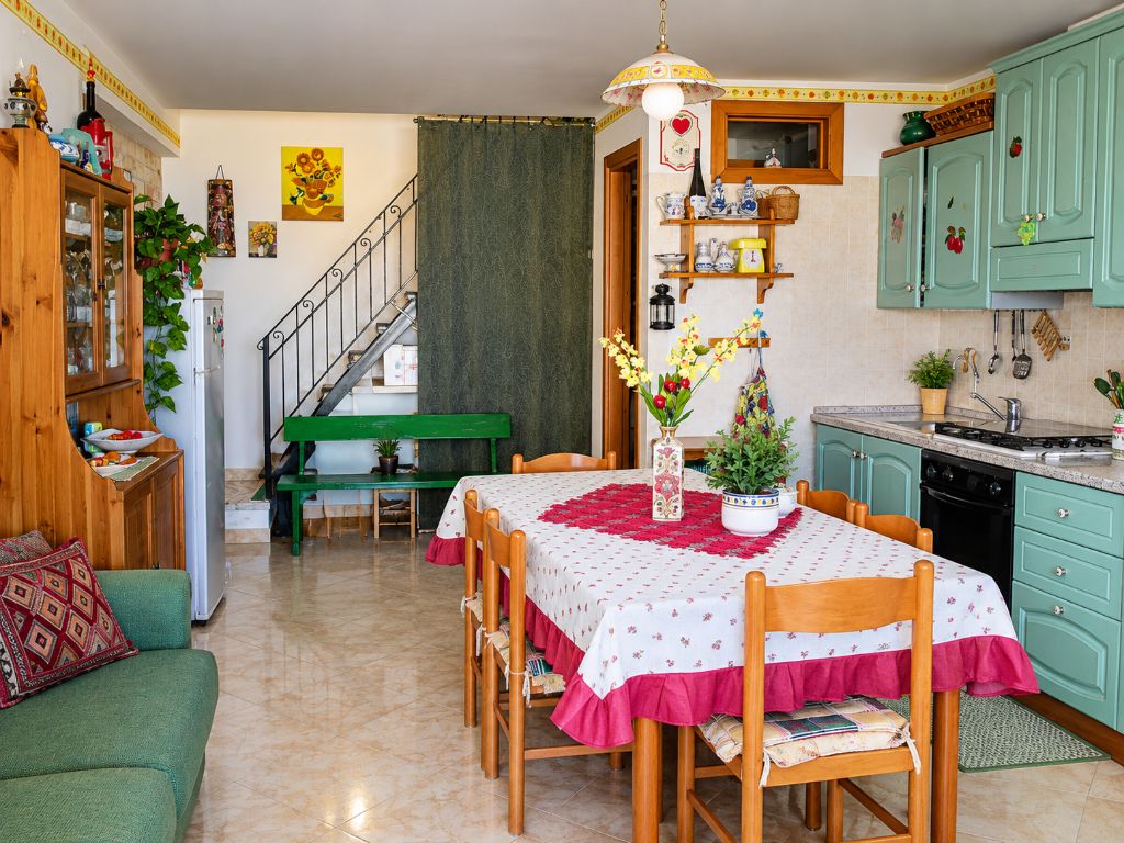 Zona pranzo con tavolo apparecchiato per quattro, un divano verde e una cucina con mobili color petrolio.