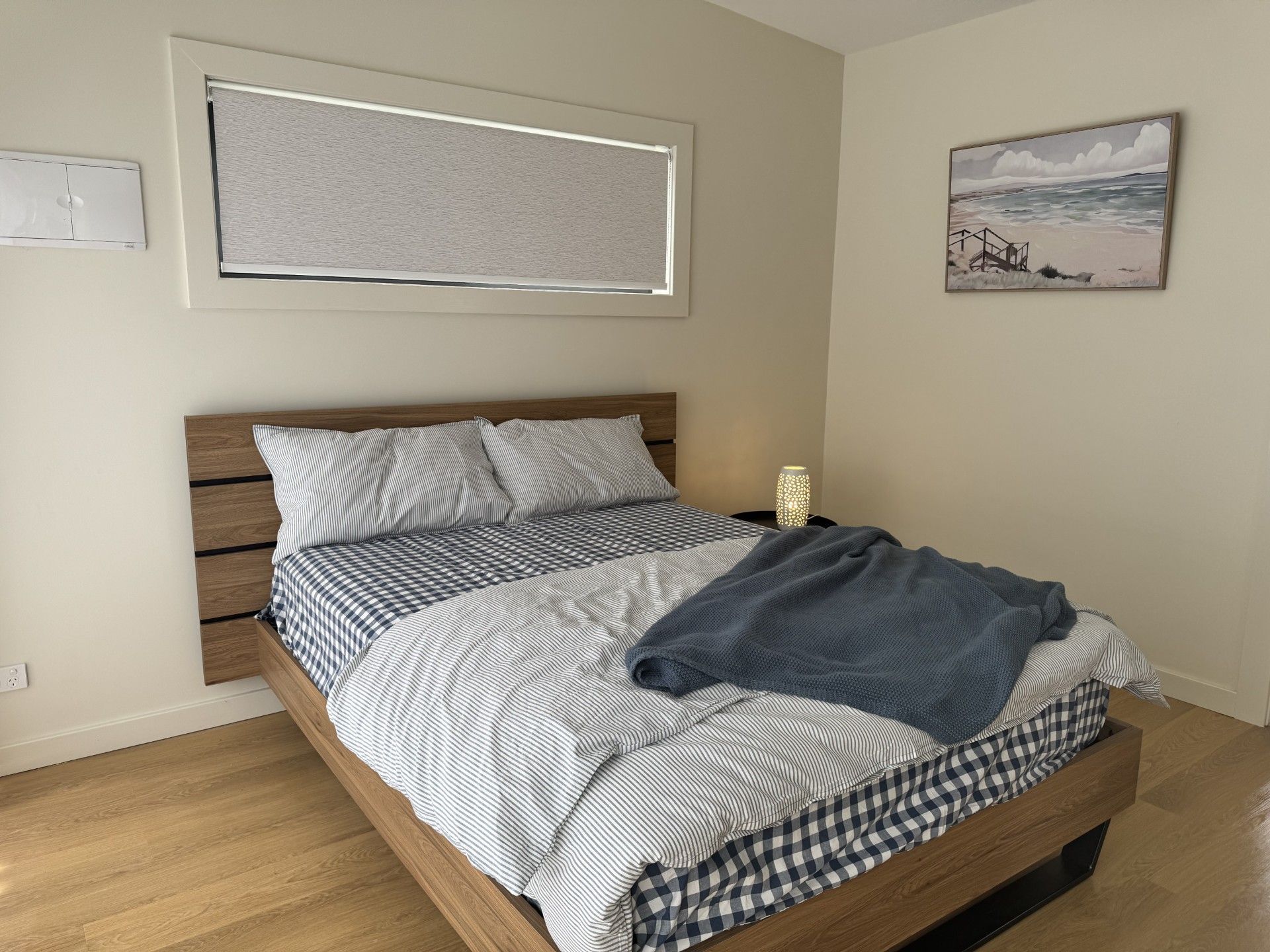 Una cama bien hecha con una colcha a rayas y almohadas, una lámpara de noche y un cuadro de una playa en la pared.
