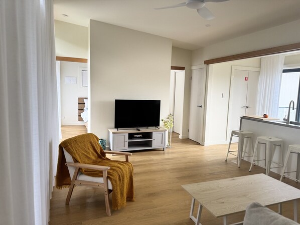 A modern living room with a flat-screen TV, a sofa, a coffee table, and a dining area with bar stools.
