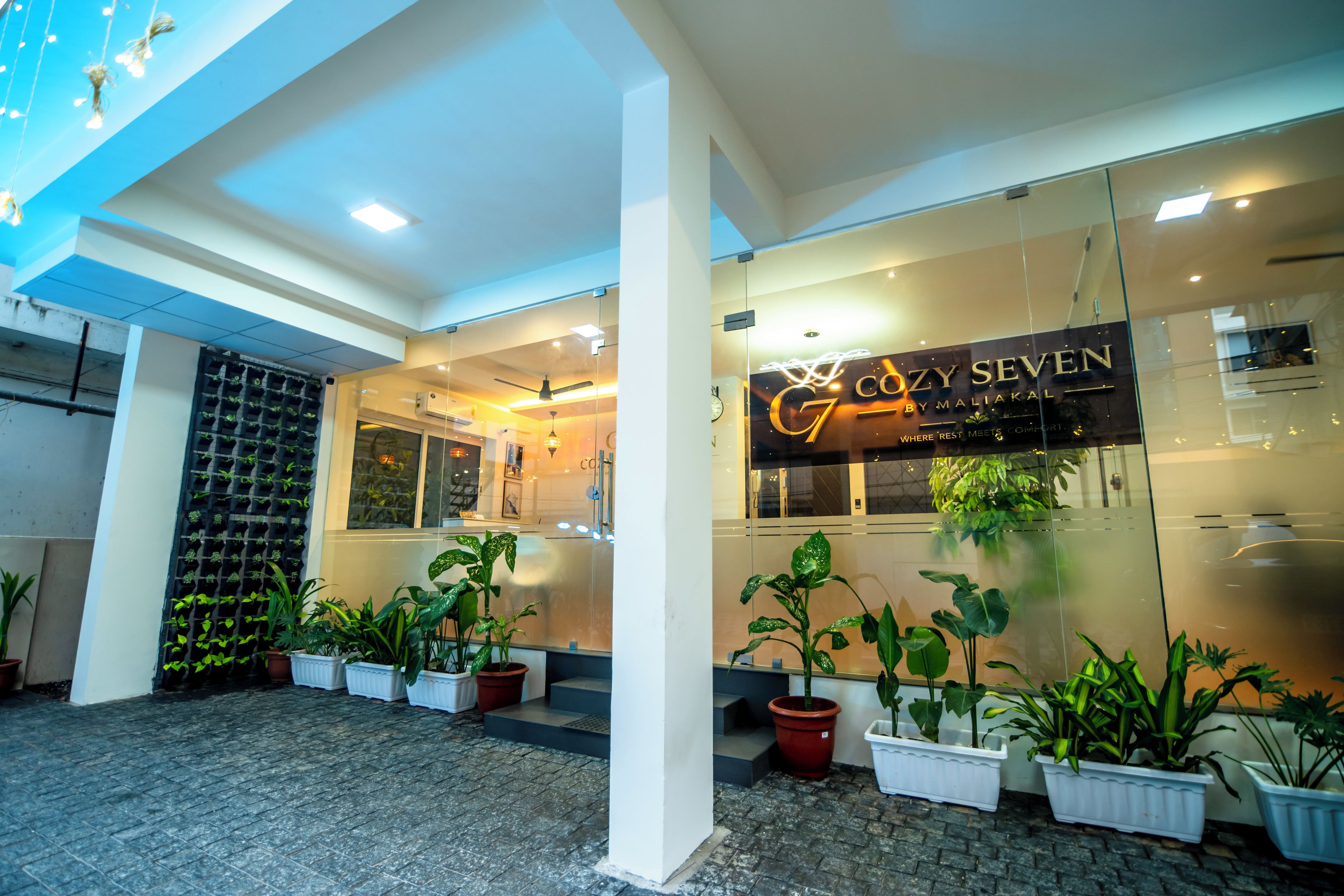 Entrada de hotel con fachada de vidrio, plantas en macetas y un letrero que dice 'COZY SEVEN'.