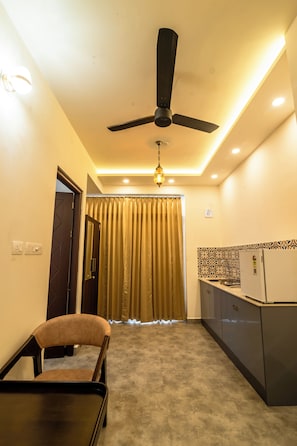 A modern living room with a ceiling fan, a dining table with a chair, a kitchen area with a refrigerator, and a window with curtains. - Cozy Seven - 100m to North Railway Station (Kanayannur)