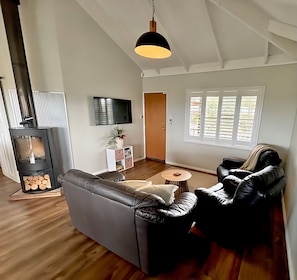 Een moderne woonkamer met een bank, lederen fauteuils, een televisie, een open haard en een raam met zonwering.