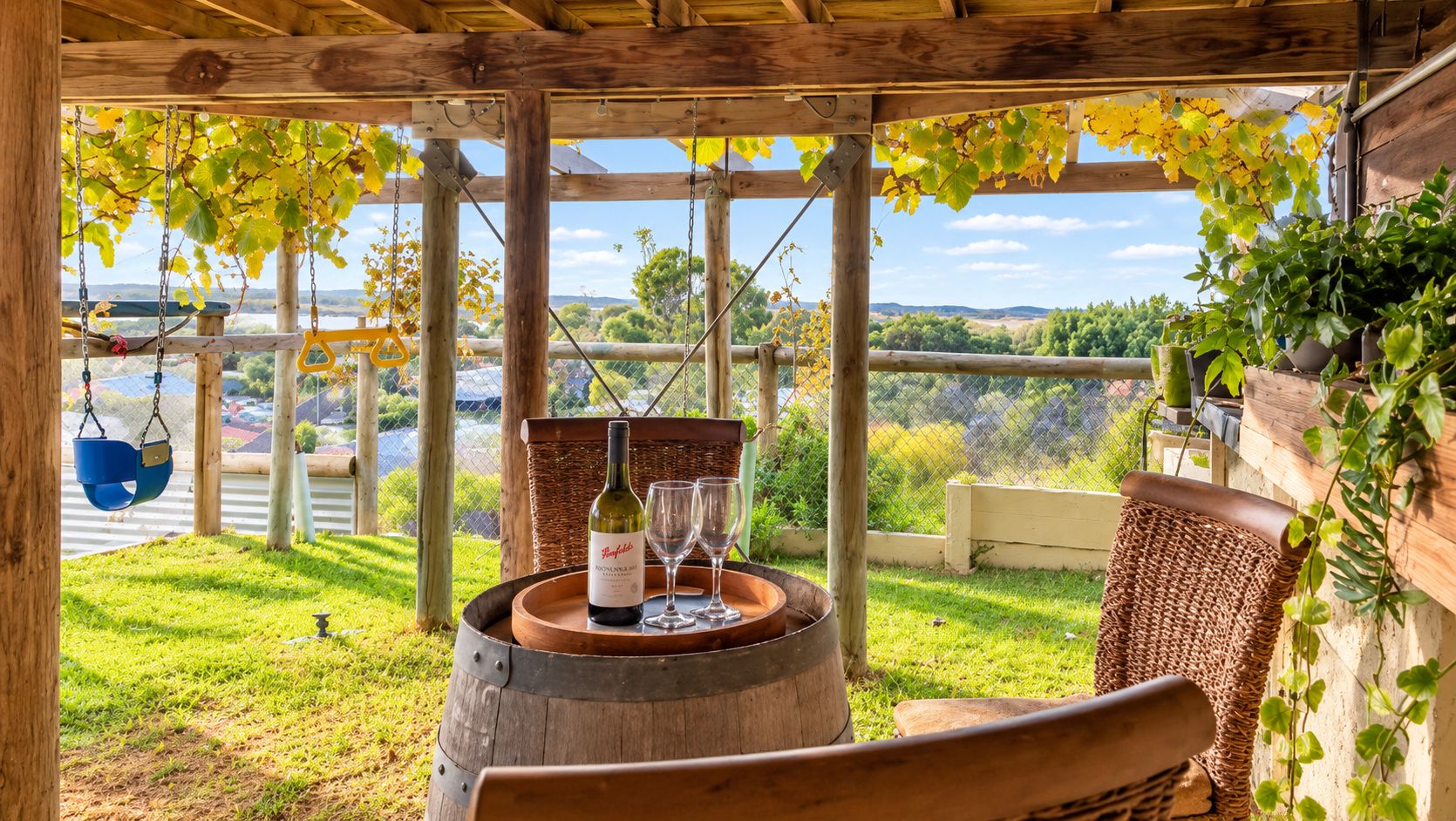 Una pérgola de madera con una mesa, una botella de vino y copas, con vista a un área verde con árboles y una cerca.