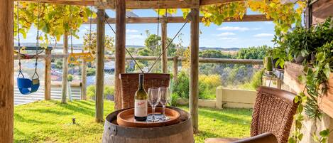 Una pérgola de madera con una mesa, una botella de vino y copas, con vista a un área verde con árboles y una cerca.