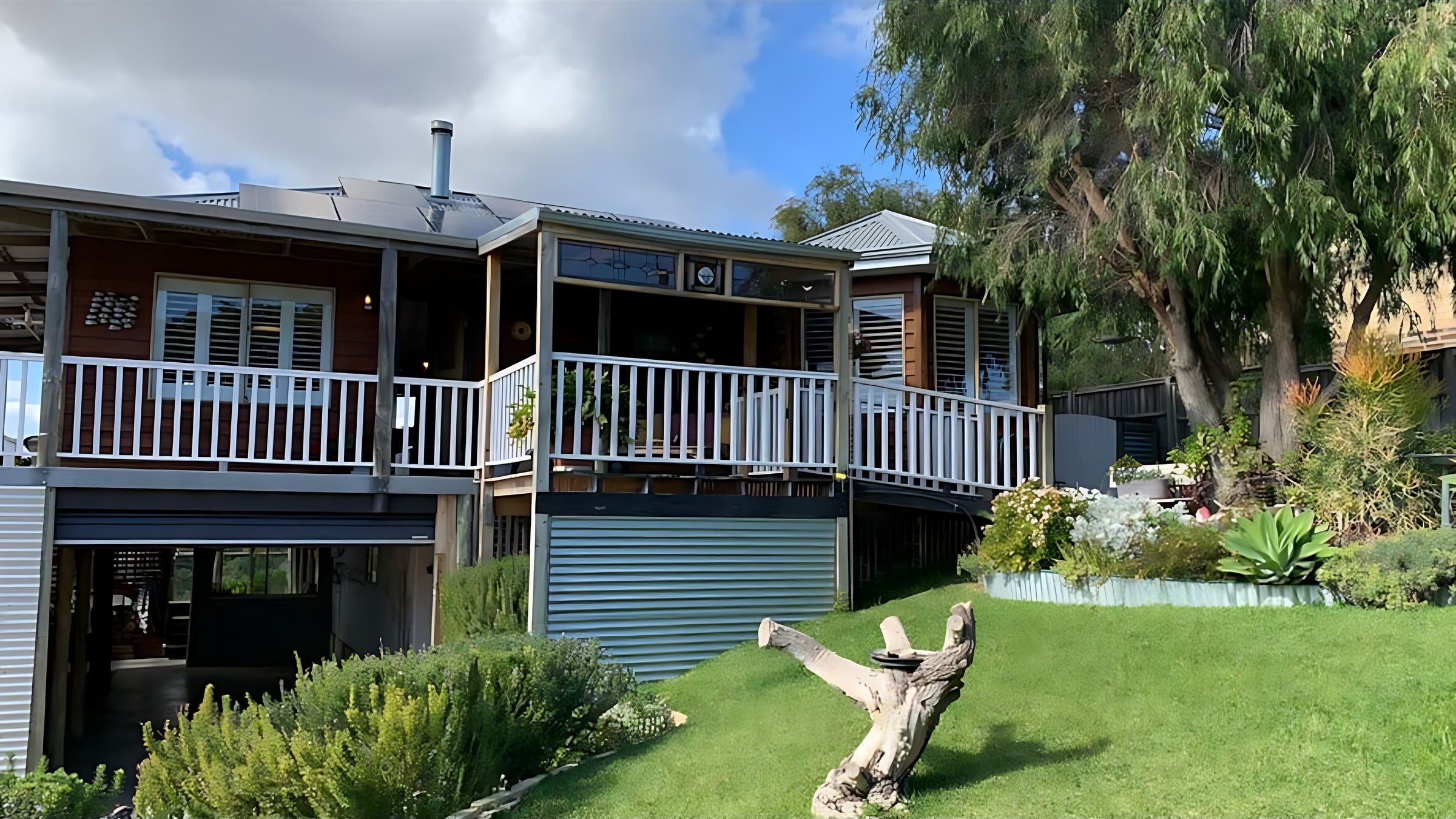 House Exterior and Veranda : Lake Views & Coastal Escape is a raised timber home with white-railed verandas, solar panels and parking underneath, set above a landscaped lawn on a quiet Preston Beach street.