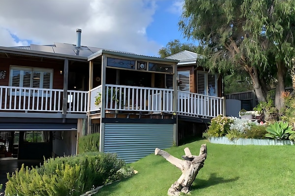 House Exterior and Veranda : Lake Views & Coastal Escape is a raised timber home with white-railed verandas, solar panels and parking underneath, set above a landscaped lawn on a quiet Preston Beach street.